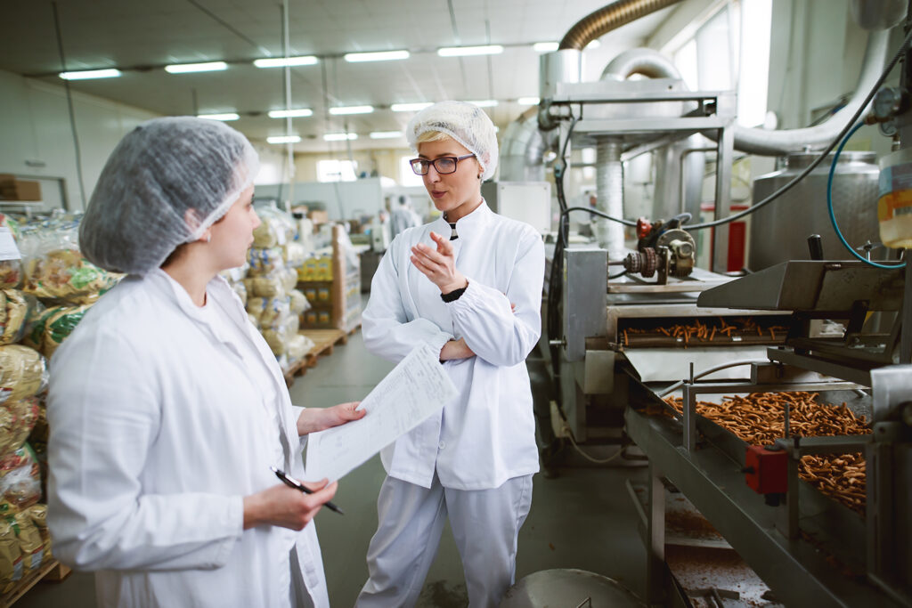 Two female workers discussing while standing in food factory. One of them holding paperwork and pen while other one speaking.