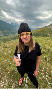 A woman in all black with yellow thick rimmed glasses and a black beanie holds a blue pen on a mountain side amidst a cloudy sky.